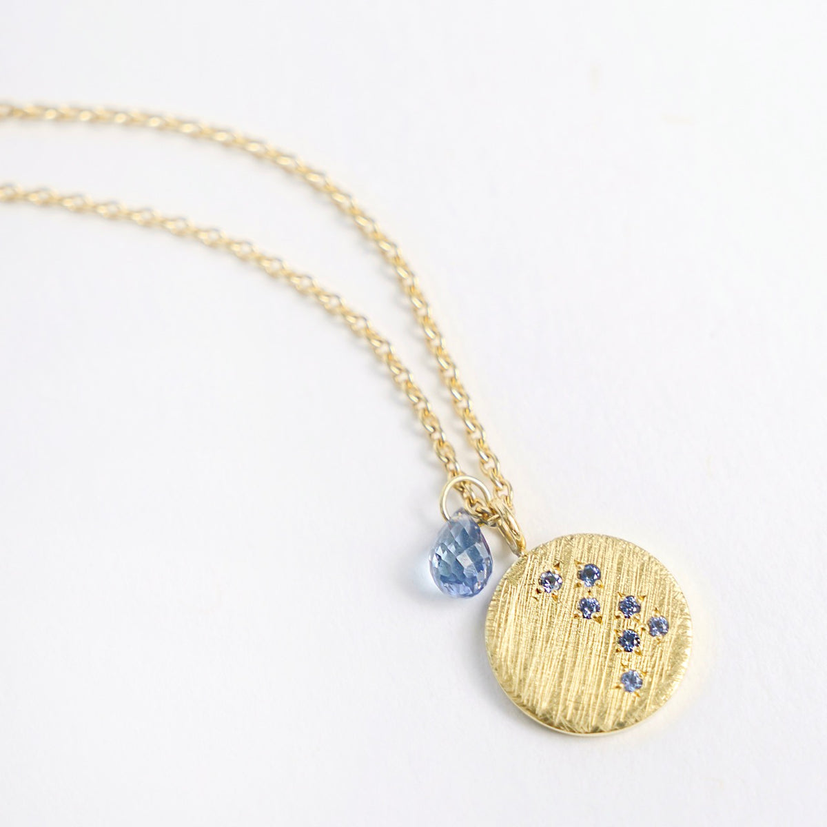 18ct yellow gold necklace featuring a textured round pendant with sapphires, and a blue teardrop charm.