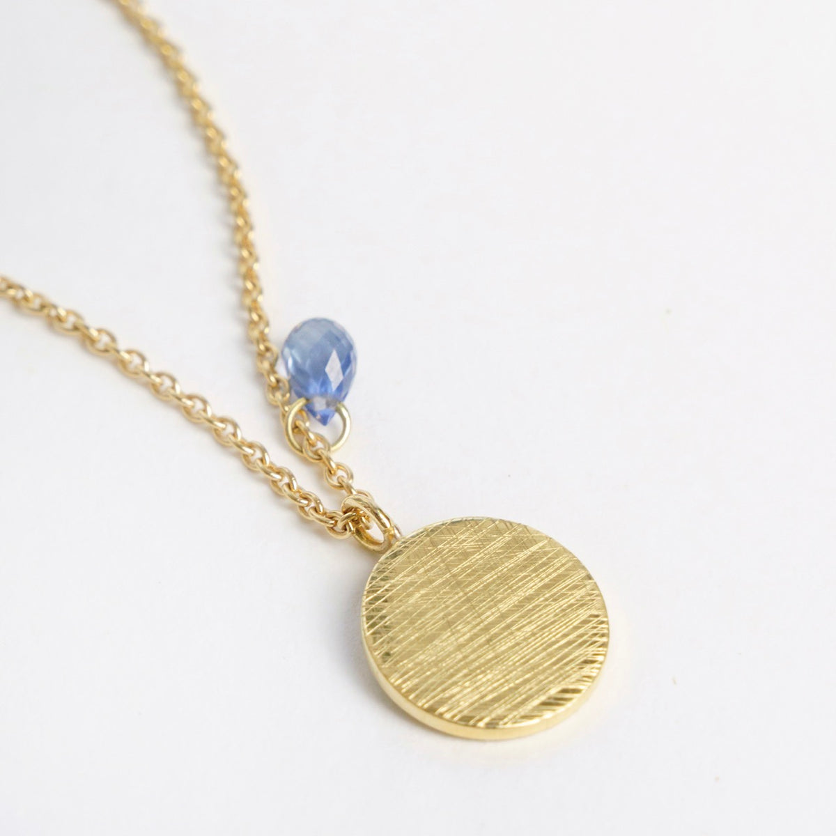 Gold necklace featuring a textured round pendant with a blue sapphire charm, set on a flat curb chain.