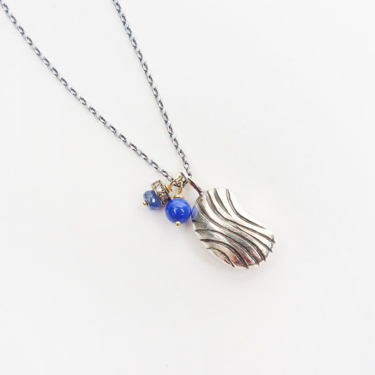 Silver bean pendant with a textured design, featuring blue lapis lazuli and sapphire beads on a blackened sterling silver chain.