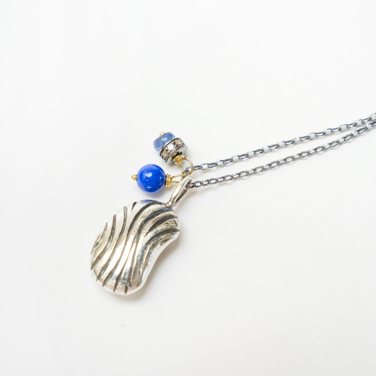 Carved Bean Pendant 2 features a hand-carved silver bean with textured grooves, accompanied by blue gemstones.