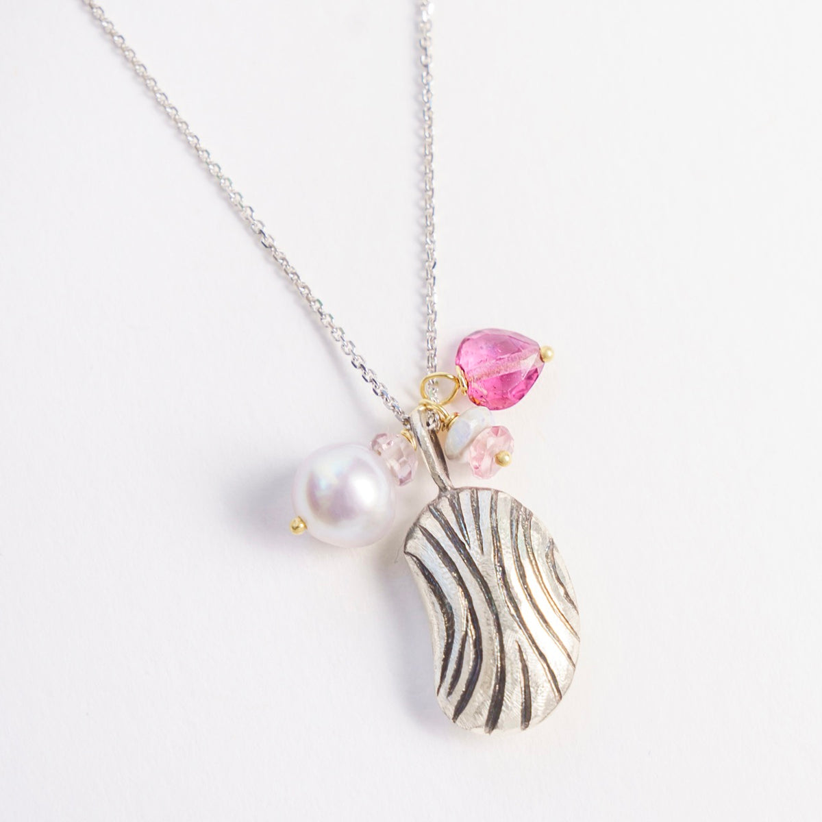 Silver bean-shaped pendant with grooves, adorned with a freshwater pearl, pink tourmaline, and other gemstones, on a chain.