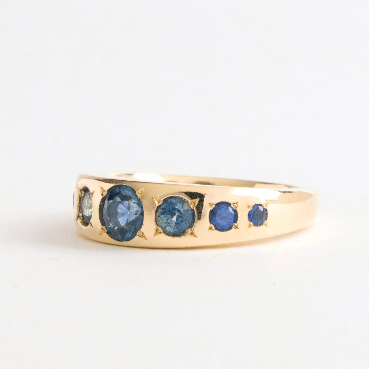 Seven Sisters Band Ring featuring a row of seven blue sapphires set in a polished 18ct yellow gold band.