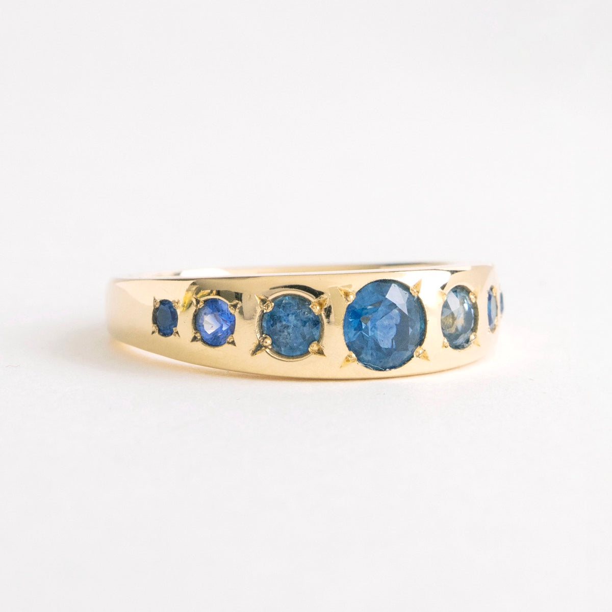 Seven Sisters Band Ring in 18ct yellow gold featuring seven blue sapphires of varying sizes and tones.