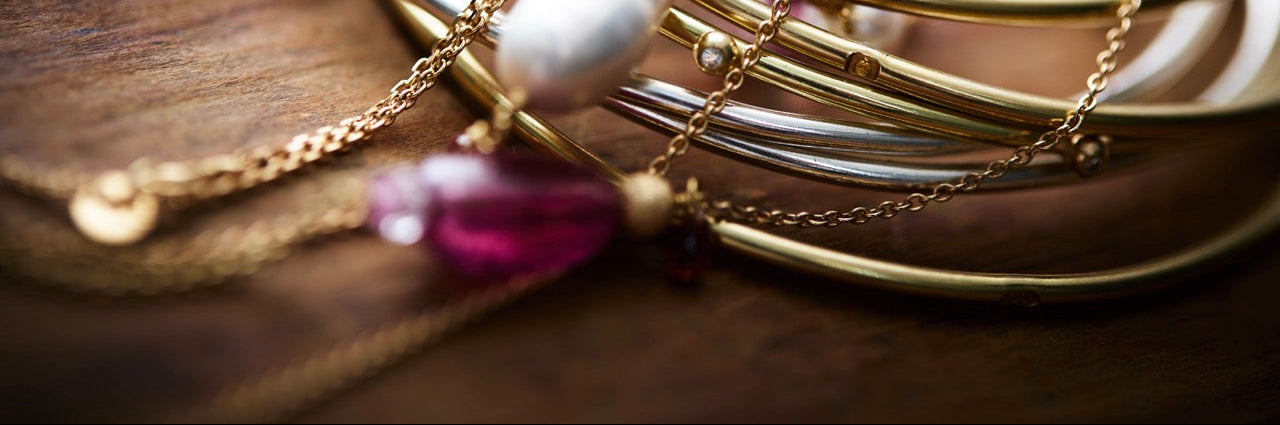 A close-up of stacked gold bangle bracelets with layered chain straps, a white pearl-like bead, and pink gemstone accents, arranged on a dark wooden surface.